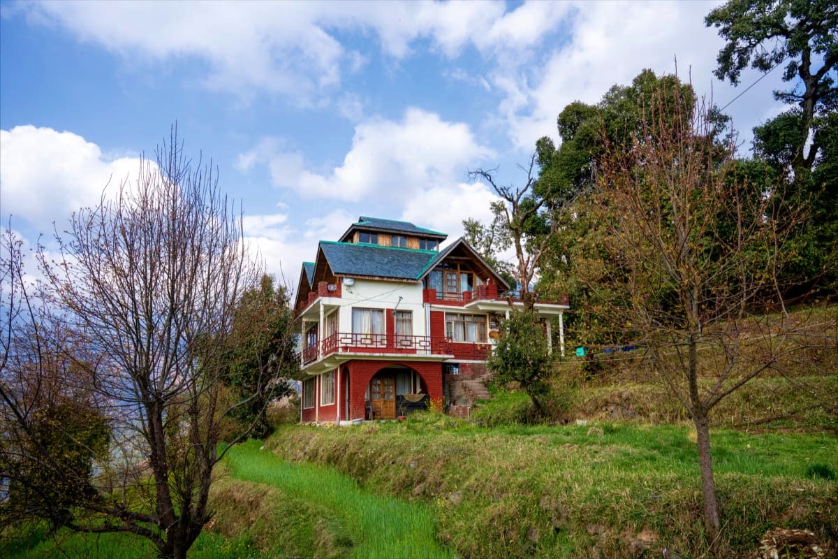 Historic red and white Victorian mansion on hillside with green shutters and turquoise roof, surrounded by bare trees and gre