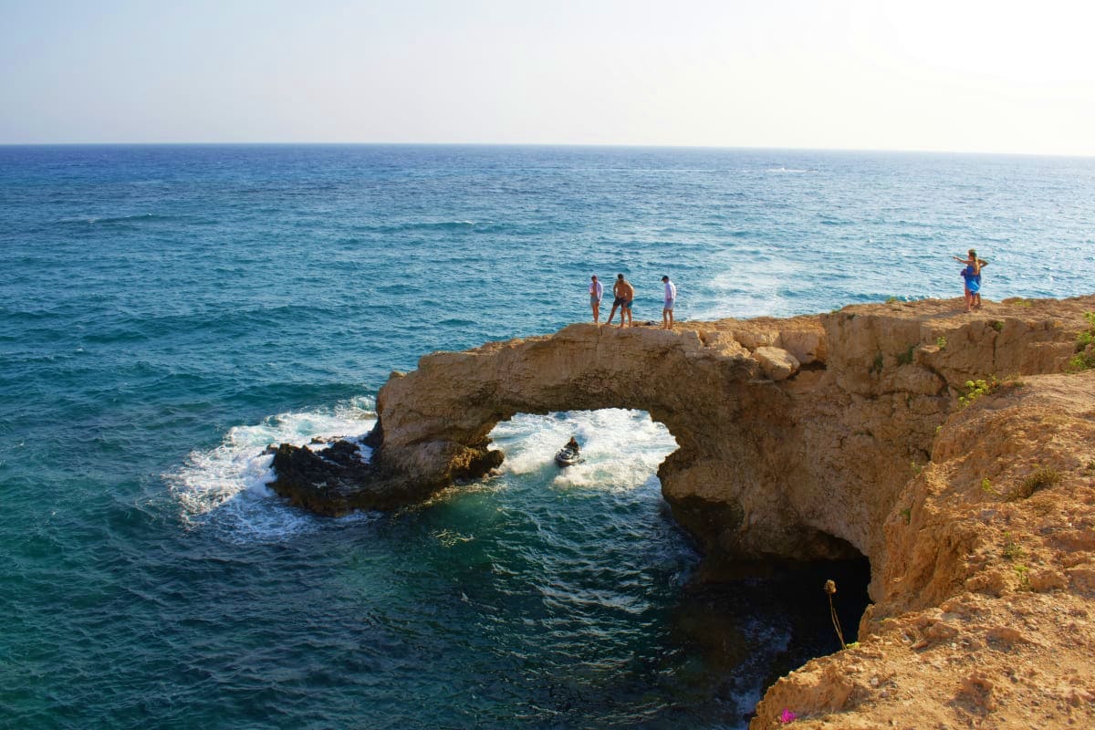 Tourists standing on rocky cliff with natural stone arch formation overlooking Mediterranean Sea in Cyprus