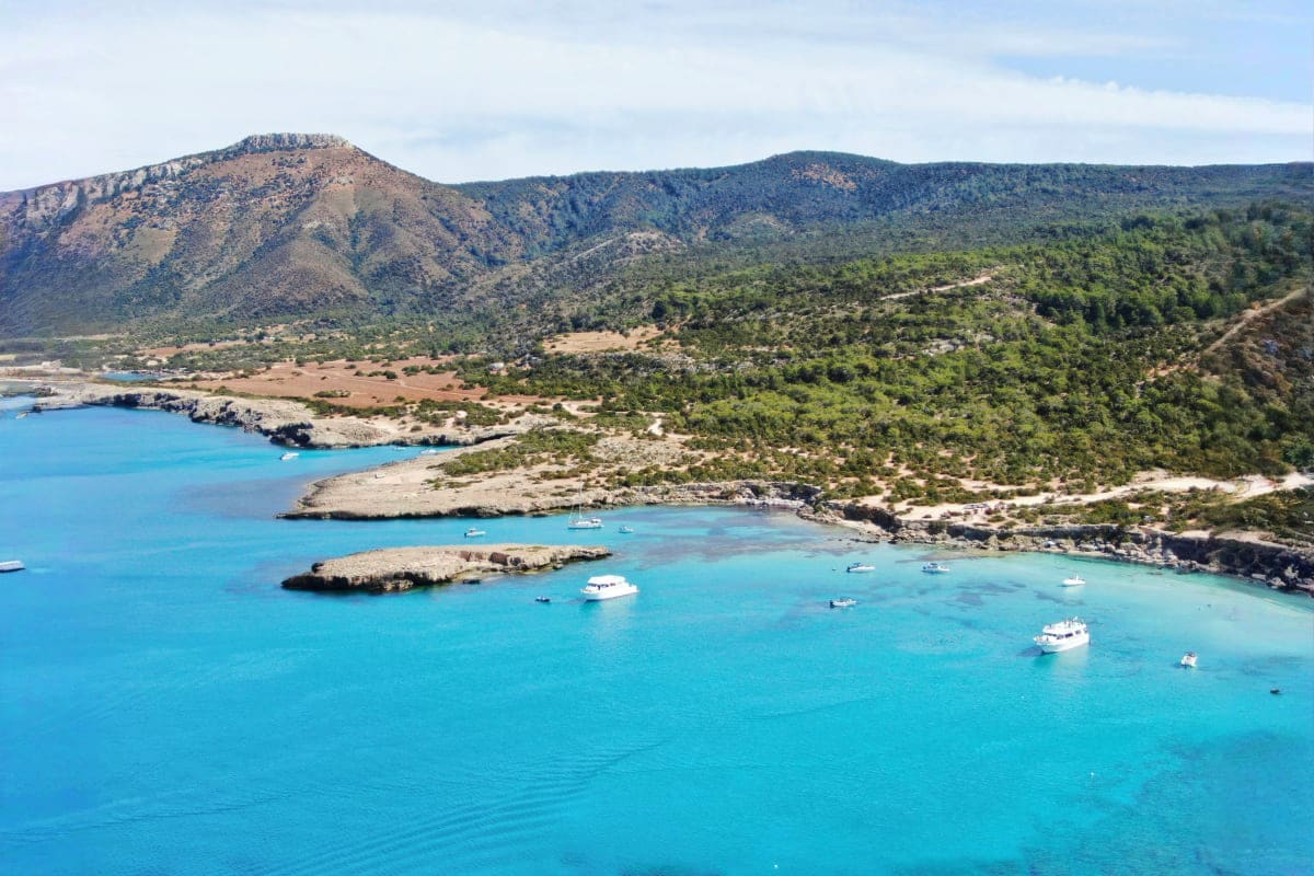 Turquoise Mediterranean cove with anchored boats, rocky coastline, and forested mountains under blue sky in Cyprus
