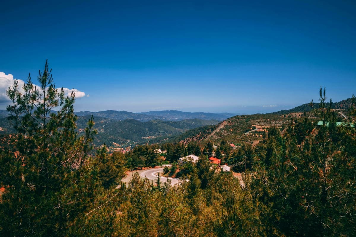 Mountainous landscape of Cyprus with winding road, scattered houses, pine forests, and distant valleys under clear blue sky