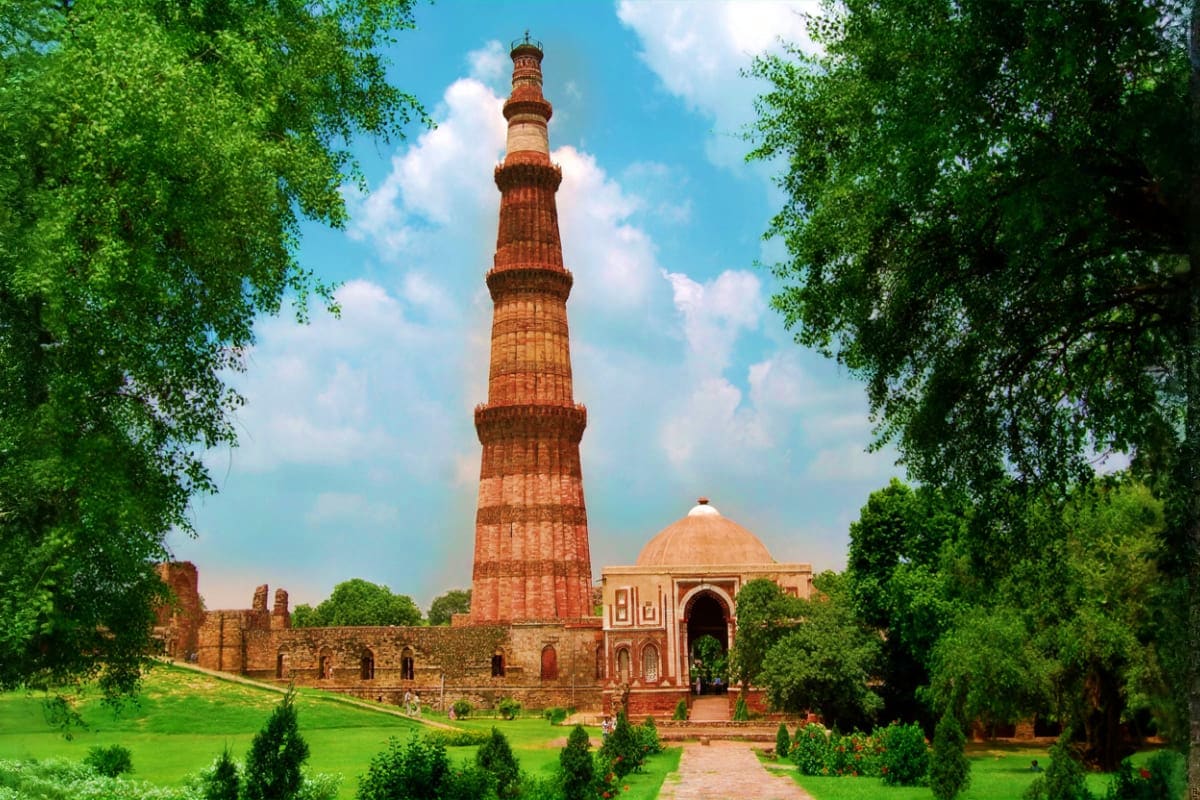 Qutb Minar monument in Delhi surrounded by lush green gardens and historic ruins with blue sky and clouds