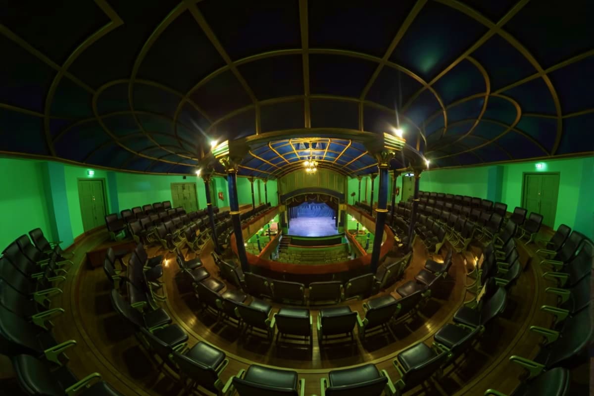 Historic theater interior with ornate gold ceiling dome, rows of black seats, green walls, and illuminated stage with spotlig