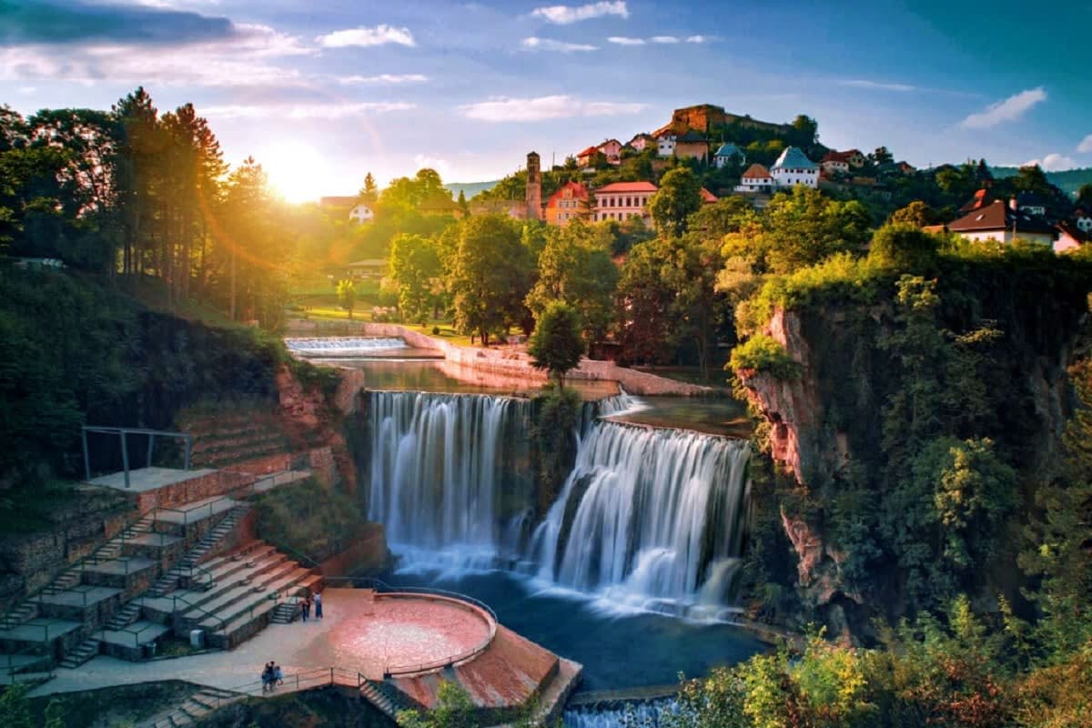 Explore Bosnia & Herzegovina Waterfall cascading over rocky cliffs in Jajce, Bosnia with historic town buildings nestled on hillside at sunset
