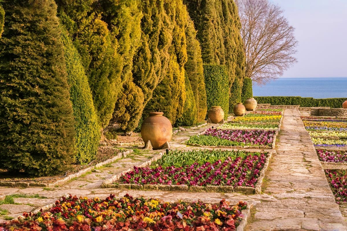 Formal Mediterranean garden with tall cypress trees, terracotta urns, and colorful flower beds overlooking the sea