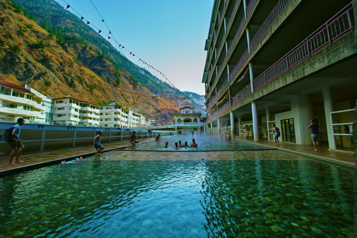 Children and adults swimming in a natural hot spring pool surrounded by modern apartment buildings and mountains in Manali