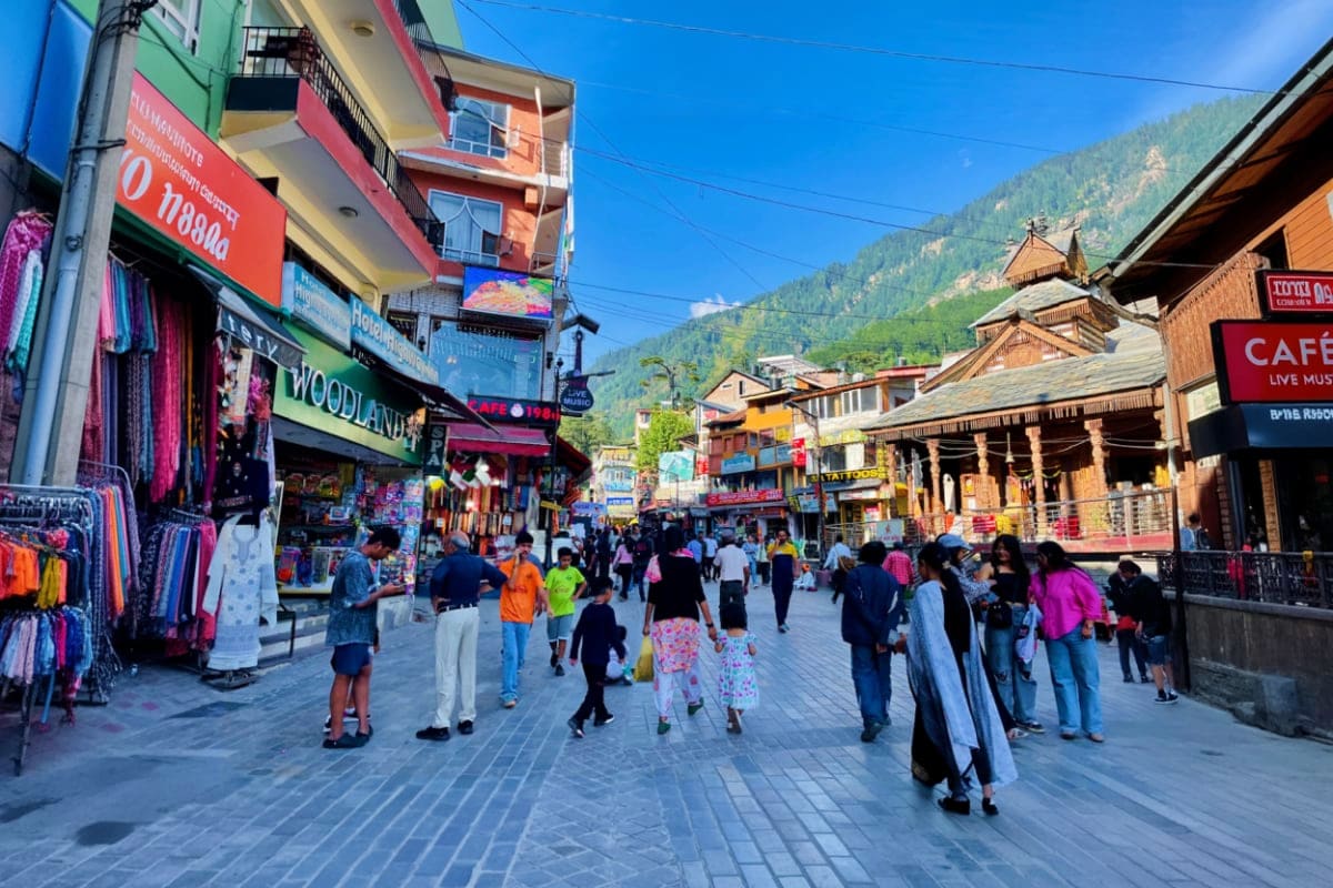 Crowded pedestrian shopping street in Manali with colorful storefronts, wooden architecture, and forested mountain backdrop u