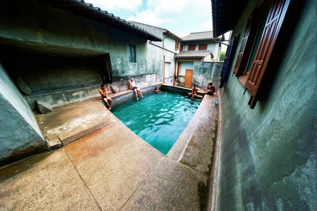 Tourists bathing in turquoise hot spring pool surrounded by concrete walls and traditional buildings in a mountain village co