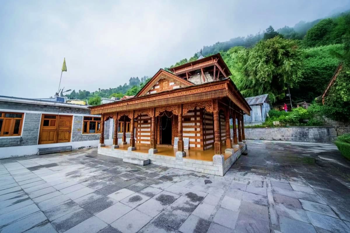 Traditional wooden temple with carved details and sloped roof in a mountain valley surrounded by green forests and stone cour