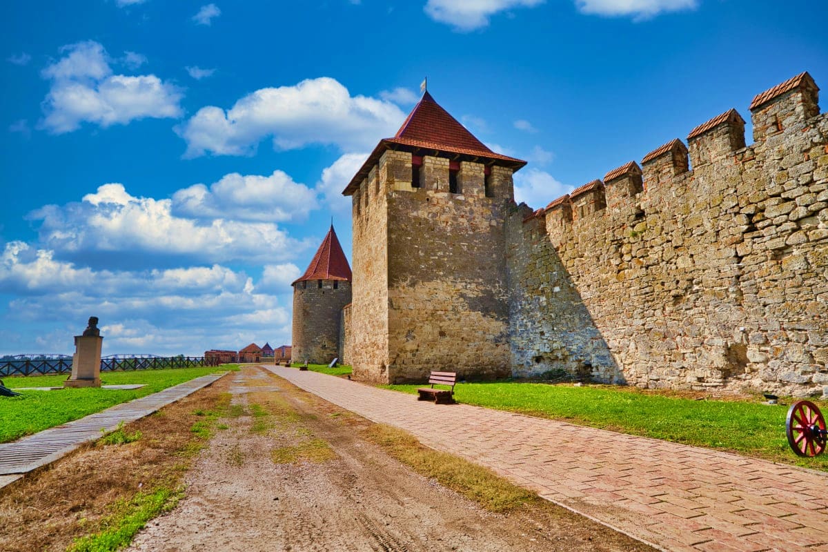 Medieval stone fortress with defensive towers and red-tiled roofs, brick pathway leading through grassy grounds under blue sk
