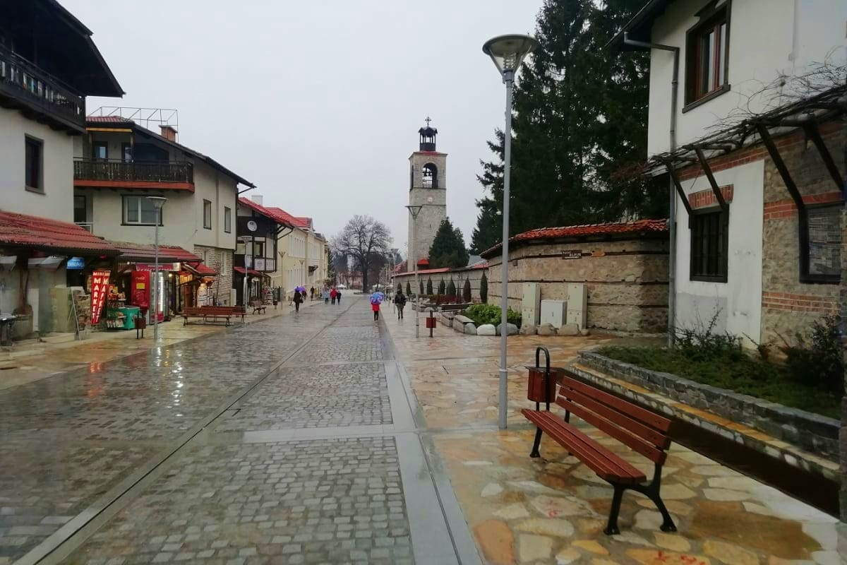 Wet cobblestone street in historic Bulgarian town square with white buildings, red-roofed houses, and church tower in backgro