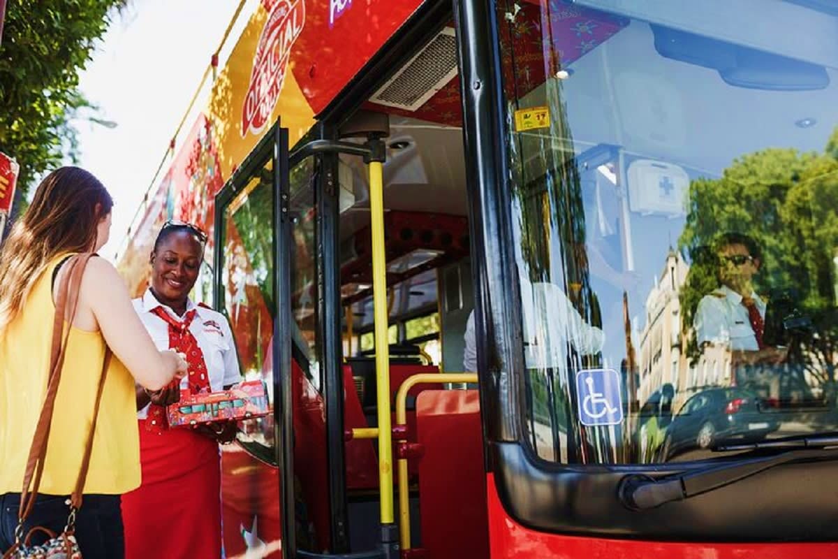 Woman boarding red and yellow tour bus with smiling attendant in traditional red uniform at Tallinn street location