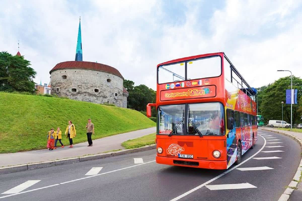 Red double-decker tour bus passing historic Tallinn tower and fortified wall with tourists on green hillside