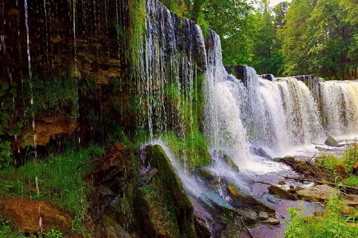 Cascading waterfall with multiple tiers flowing over moss-covered rocky cliffs surrounded by dense green forest.