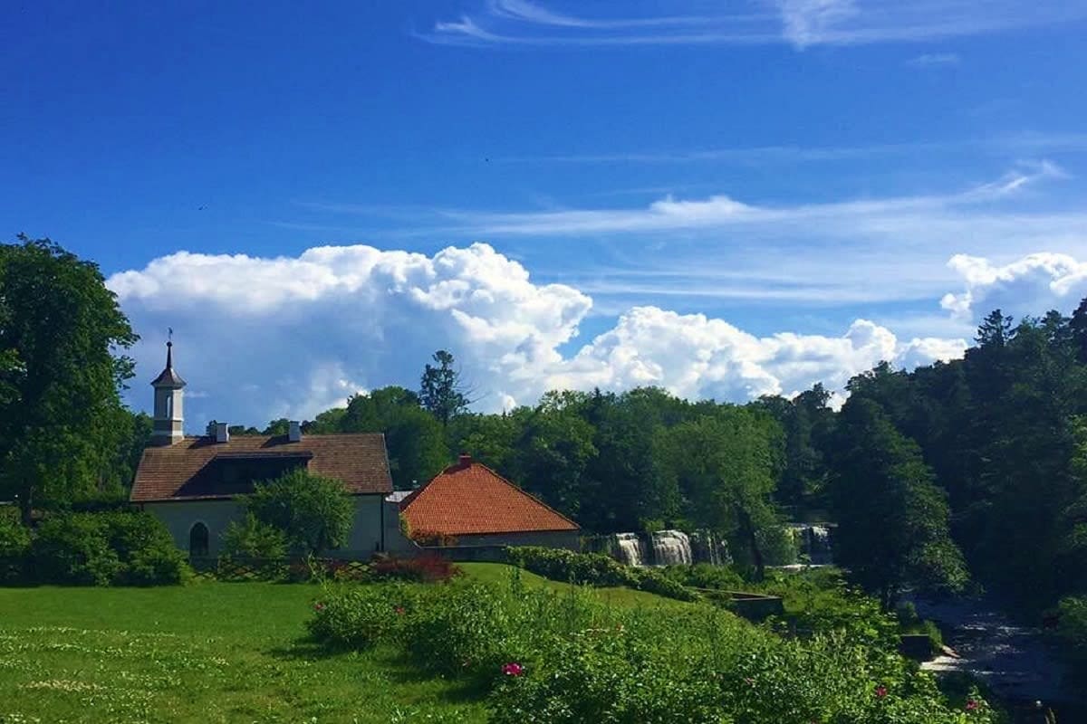 Rural Estonian countryside with white church featuring steeple, red-roofed buildings, lush green meadow, tree-lined river, an