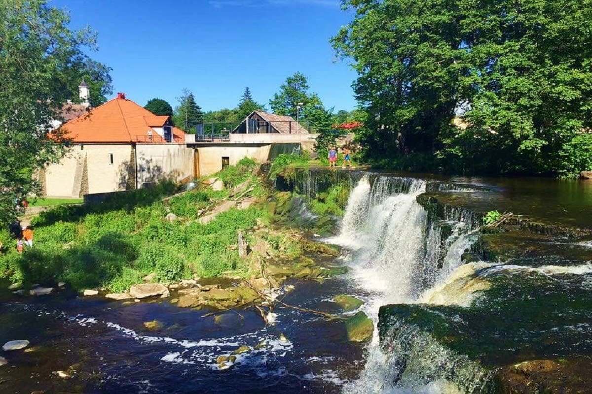 Waterfall cascading over rocky ledge beside historic cream and orange-roofed mill building surrounded by green trees and lush