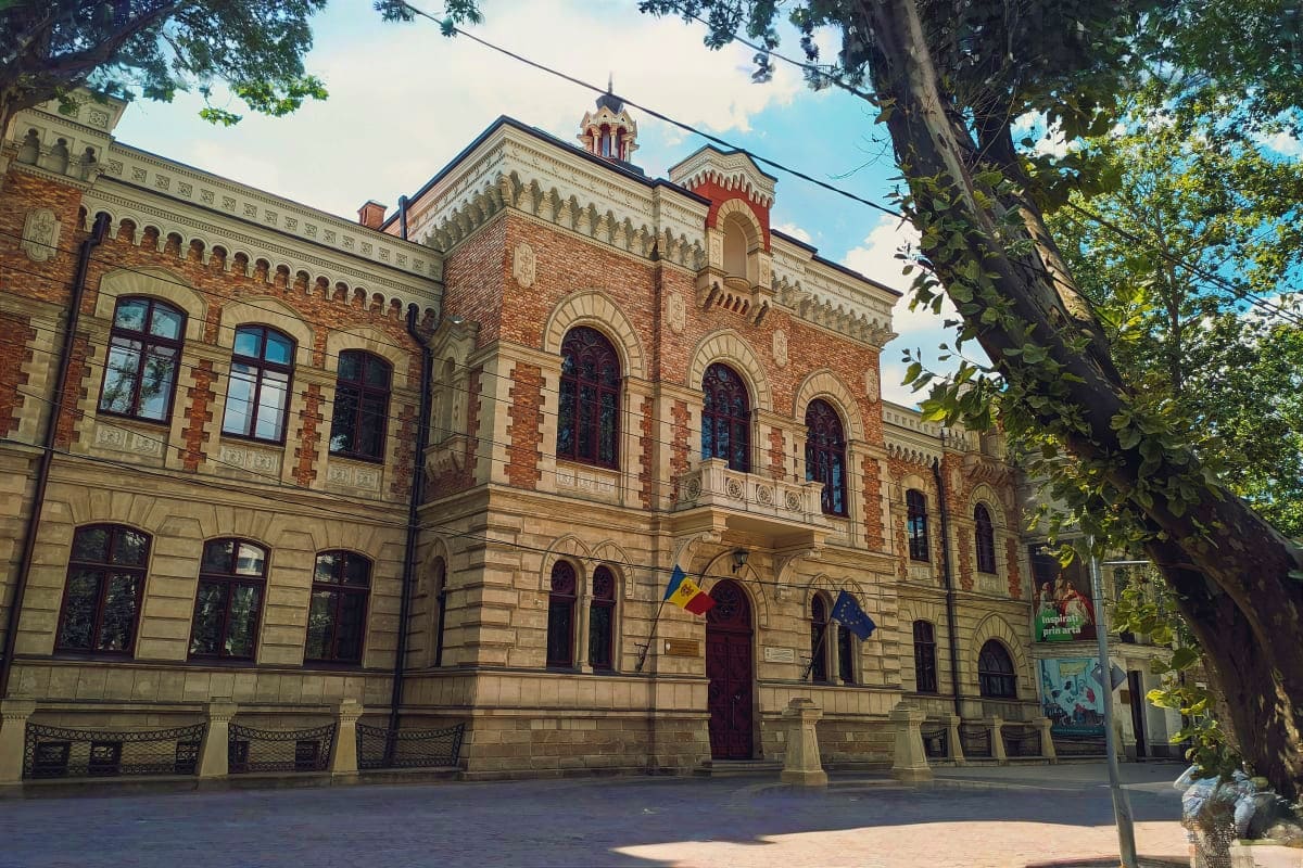 Historic brick and stone building with ornate architecture and Moldovan flag, shaded by large tree in town square