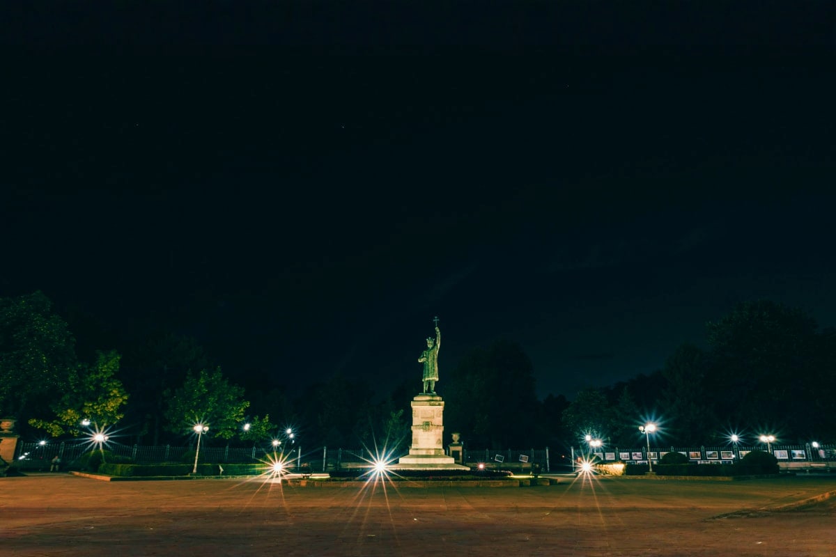Illuminated statue monument in town square at night with surrounding street lights and trees