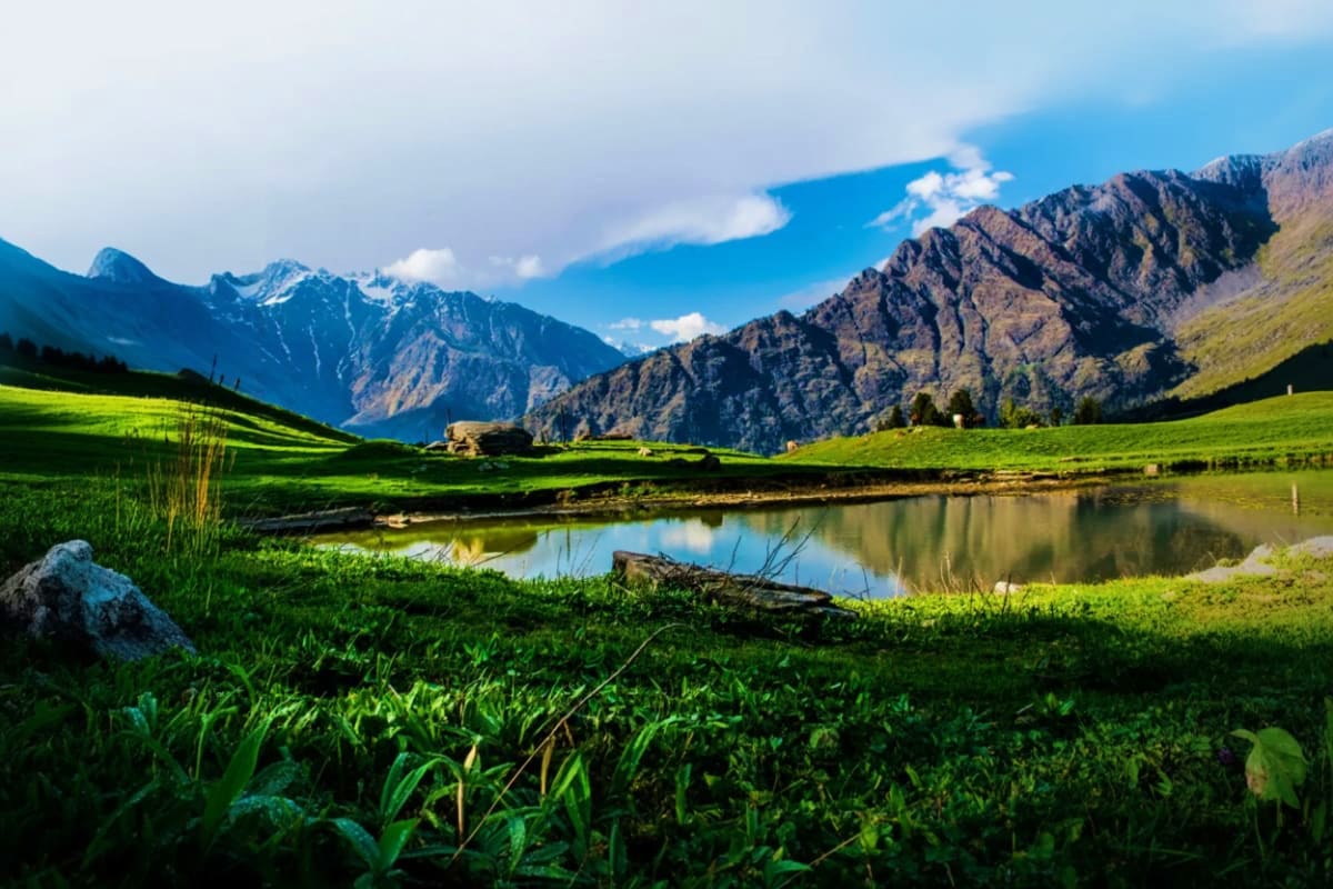 Scenic mountain valley with lush green meadows, clear stream, and snow-capped peaks under blue sky in Himalayas
