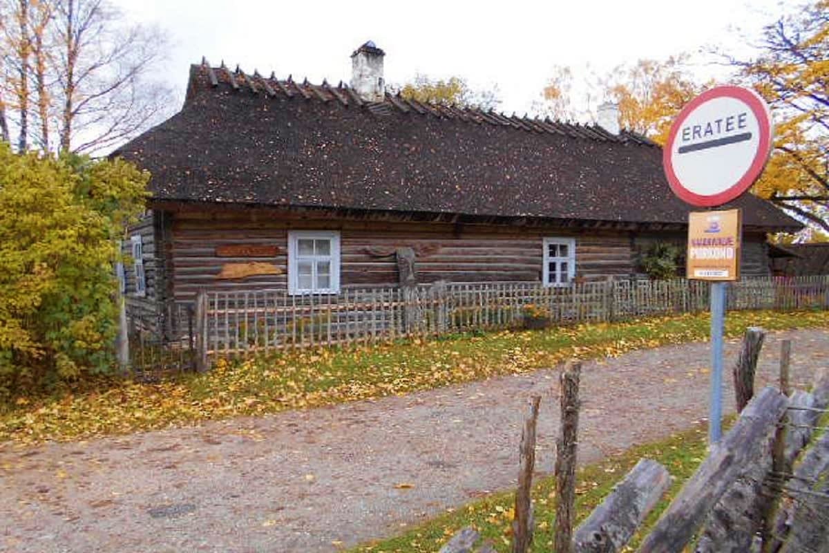Historic log cabin with dark roof and white-framed windows surrounded by autumn foliage and wooden fence in rural Estonia