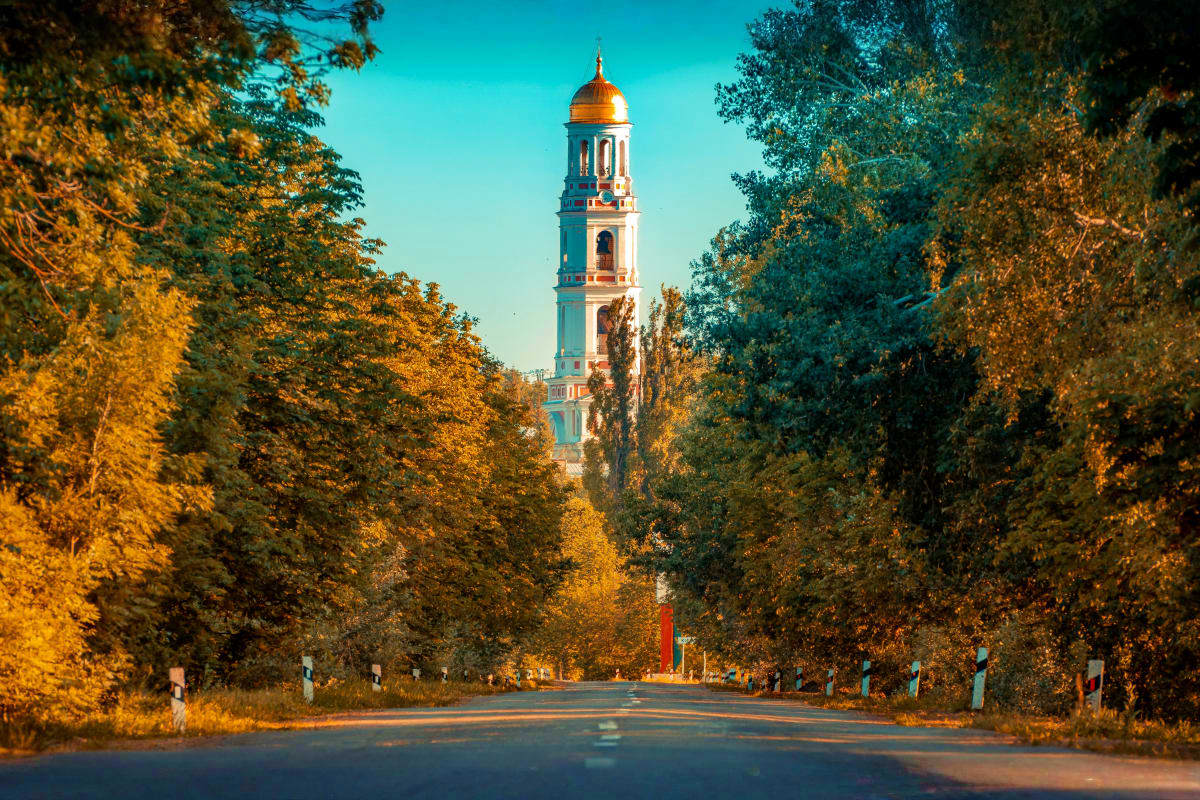 Tree-lined avenue in autumn leading to a blue and gold Orthodox church bell tower against clear turquoise sky in Moldova
