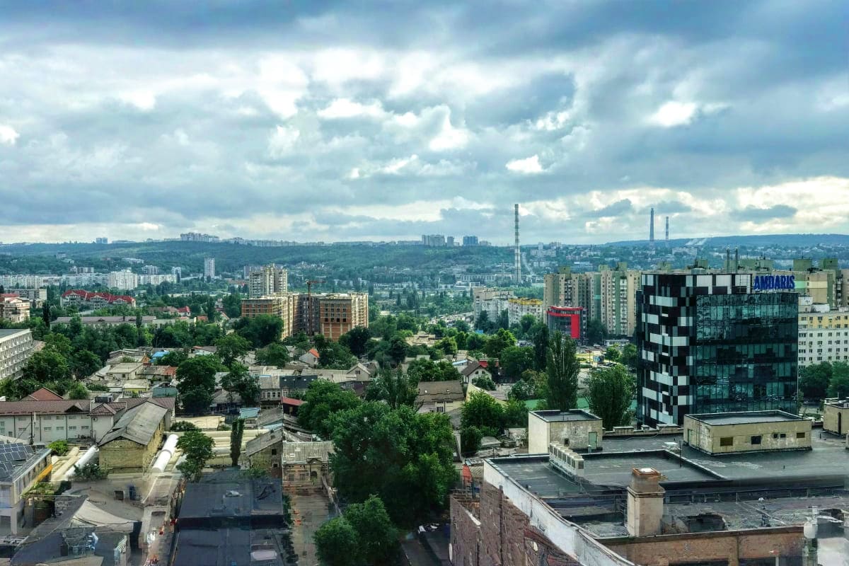 Aerial cityscape of Chisinau, Moldova showing residential buildings, green trees, and industrial smokestacks under cloudy sky