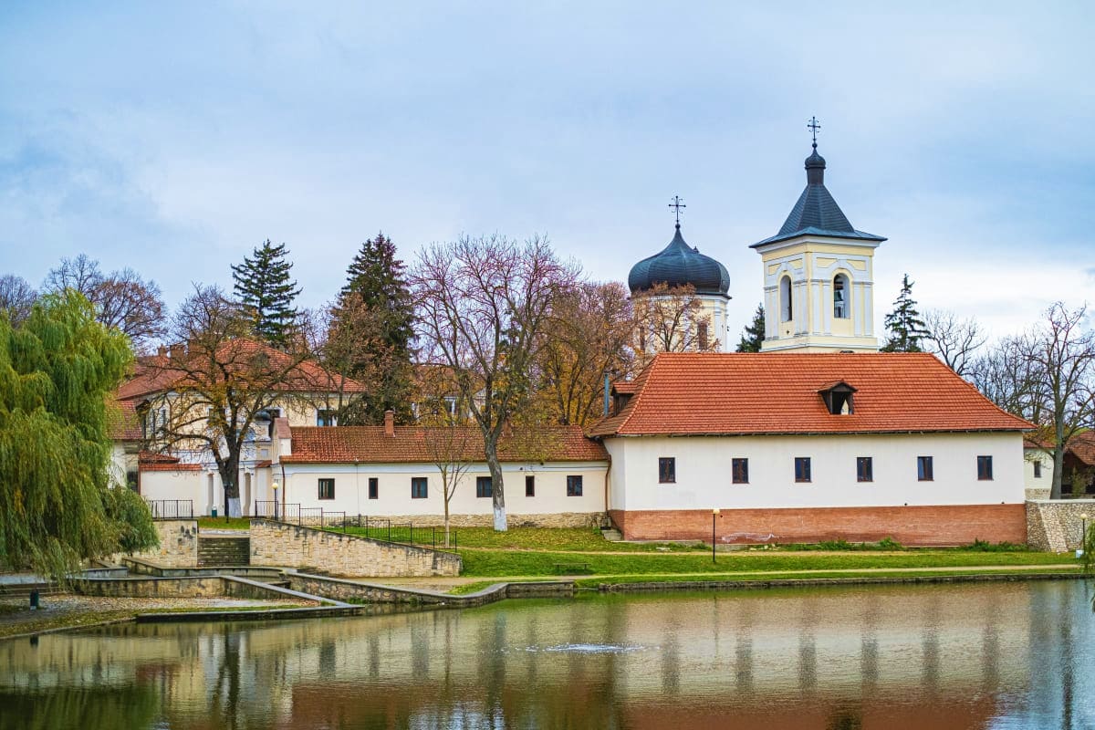 Historic Orthodox monastery with white walls and onion domes reflected in river, surrounded by bare trees in Moldova