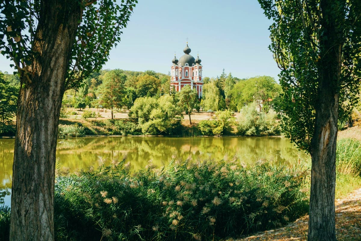 Orthodox church with golden domes reflected in river, framed by willow trees and lush green landscape in Moldova