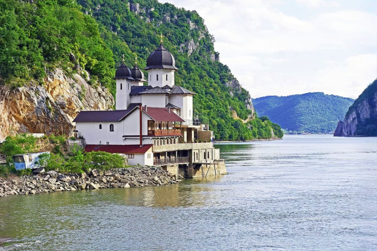Serbian Orthodox monastery with white walls and black domes on Danube River cliff surrounded by green mountains