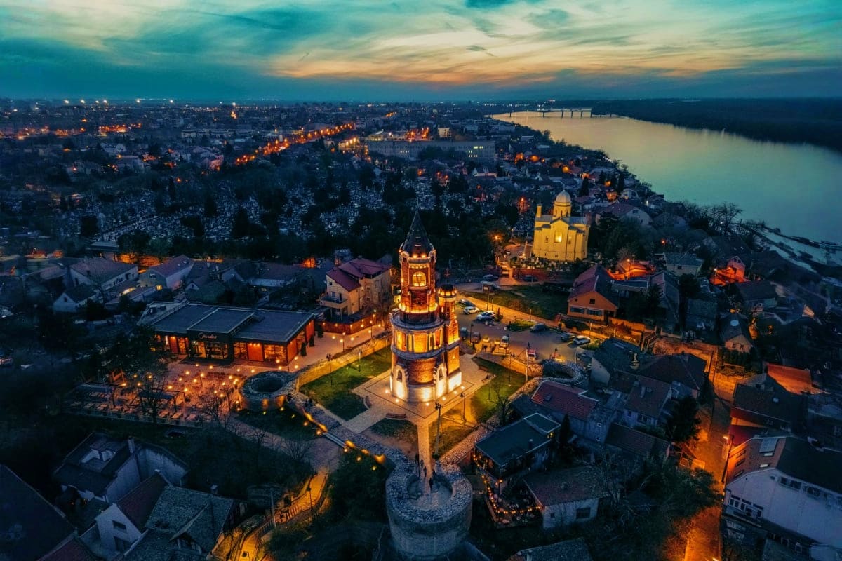 Aerial view of illuminated Orthodox churches and historic tower overlooking Danube River at dusk in Zemun, Serbia