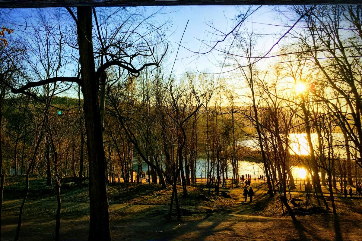 Sunset through bare trees at a riverside park with people walking along the water's edge