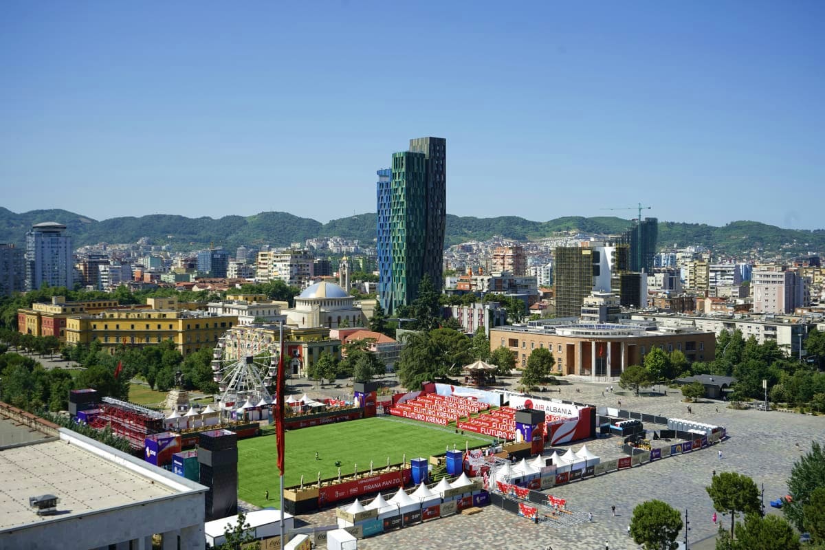 Aerial view of Tirana's main square with modern skyscraper, historic buildings, mountains, and outdoor event venue with red b