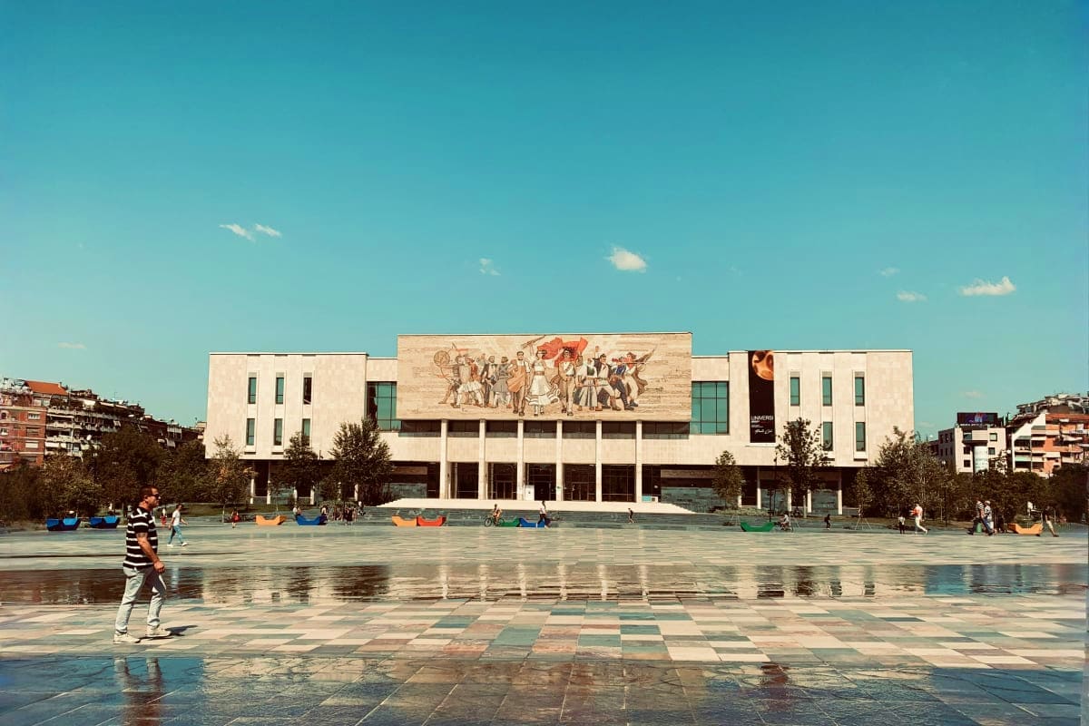 Modern museum building with classical relief sculpture on facade, public plaza with water features and visitors, clear blue s