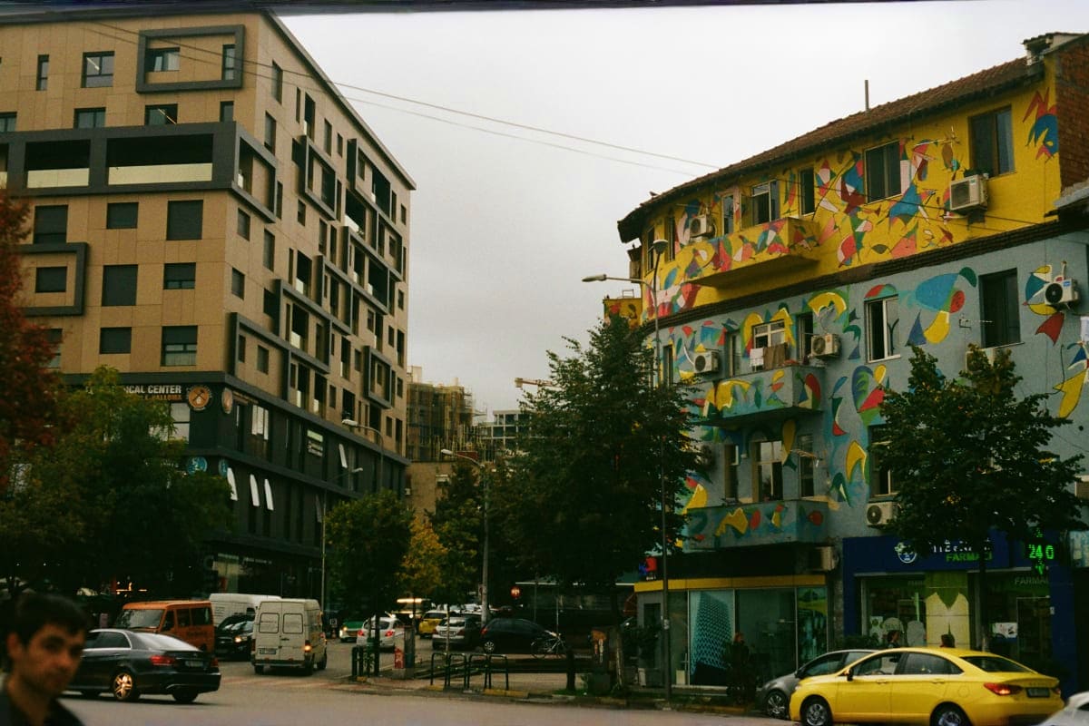 Urban street scene in Albania with colorful street art mural on yellow building, modern apartment blocks, parked cars, and ov