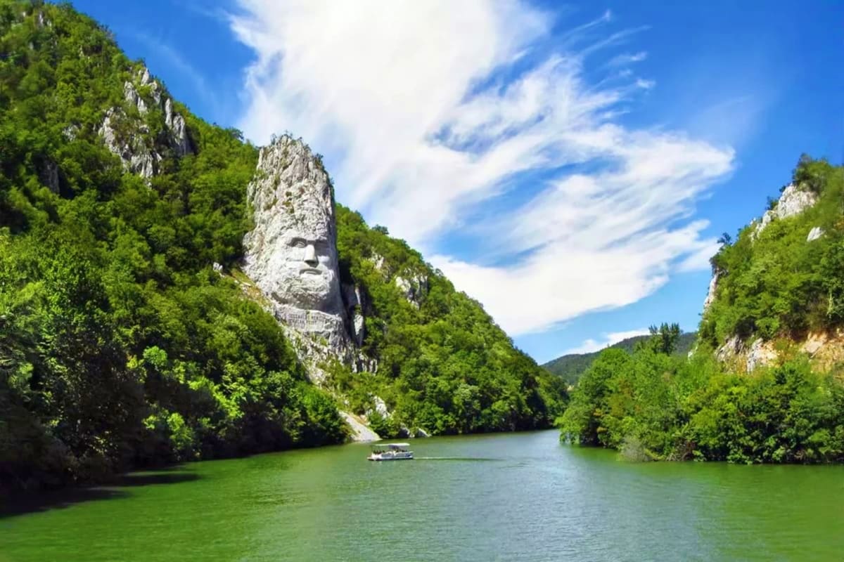 Danube River gorge with carved stone face sculpture on limestone cliff surrounded by green forest and tour boat