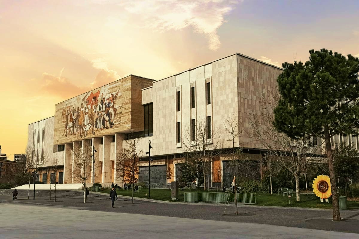 Modern museum building with stone facade and mural artwork, surrounded by plaza with trees and visitors at sunset