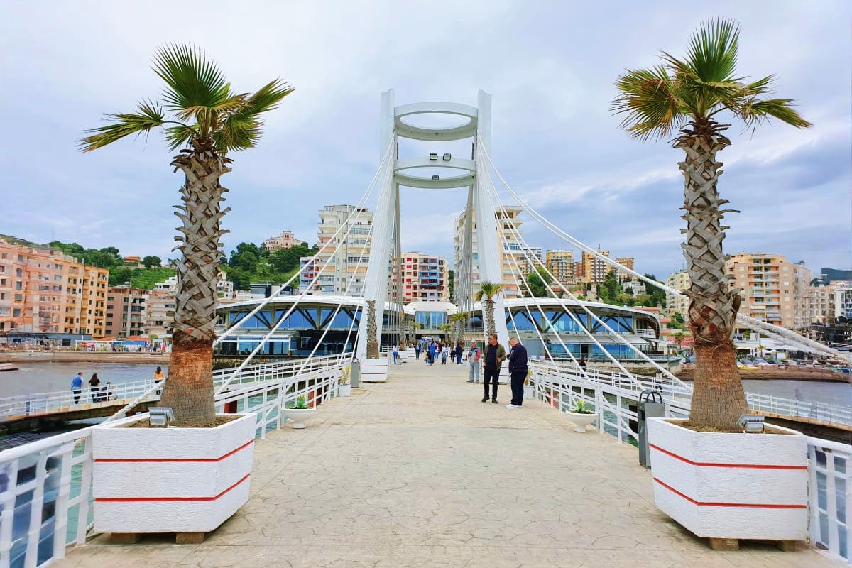 Modern white footbridge with cable stays crossing a beach, flanked by palm trees, with Mediterranean coastal city buildings i