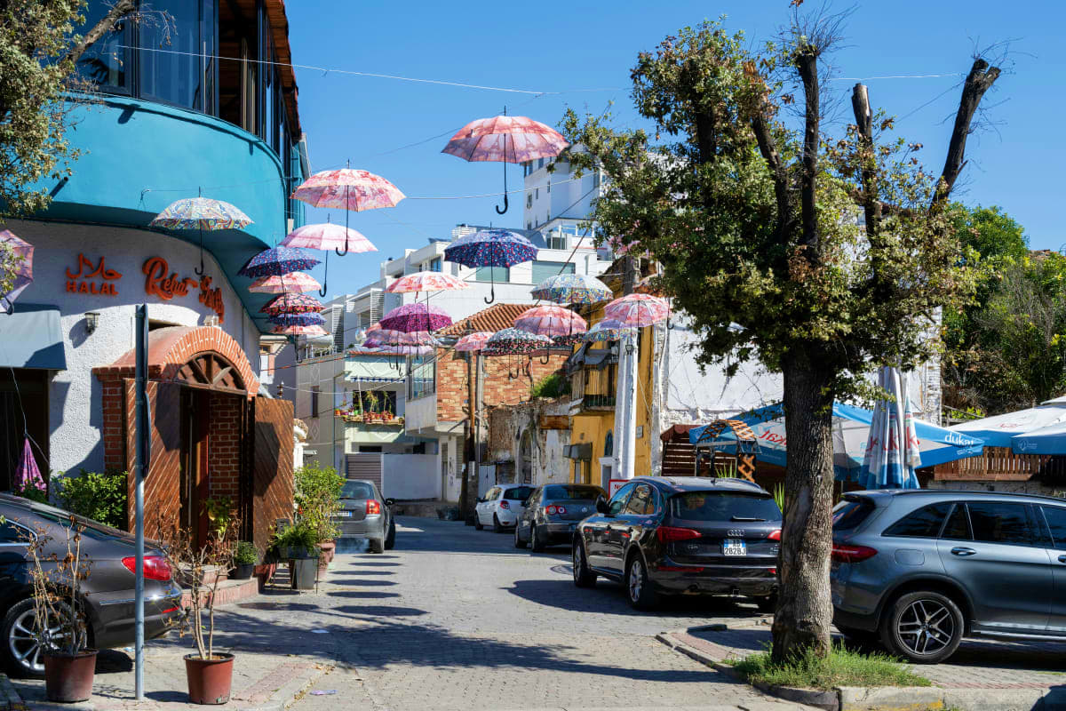 Colorful umbrellas hanging above a sunny Mediterranean street with white buildings, parked cars, and trees in Albania
