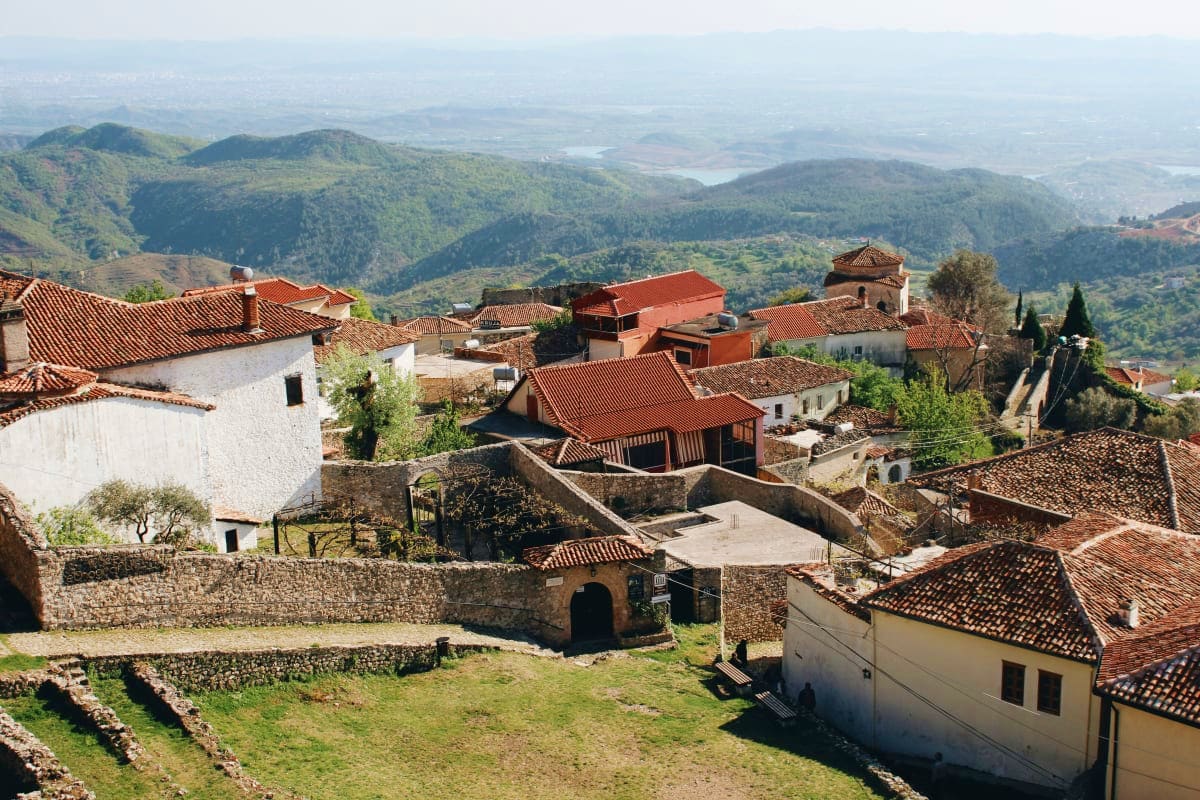Historic Albanian village with terracotta-roofed houses, stone ruins, and forested mountains in the background landscape