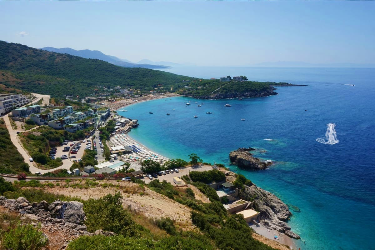Aerial view of a turquoise bay in Albania with sandy beach, anchored boats, rocky coastline, forested hillside, and waterfron
