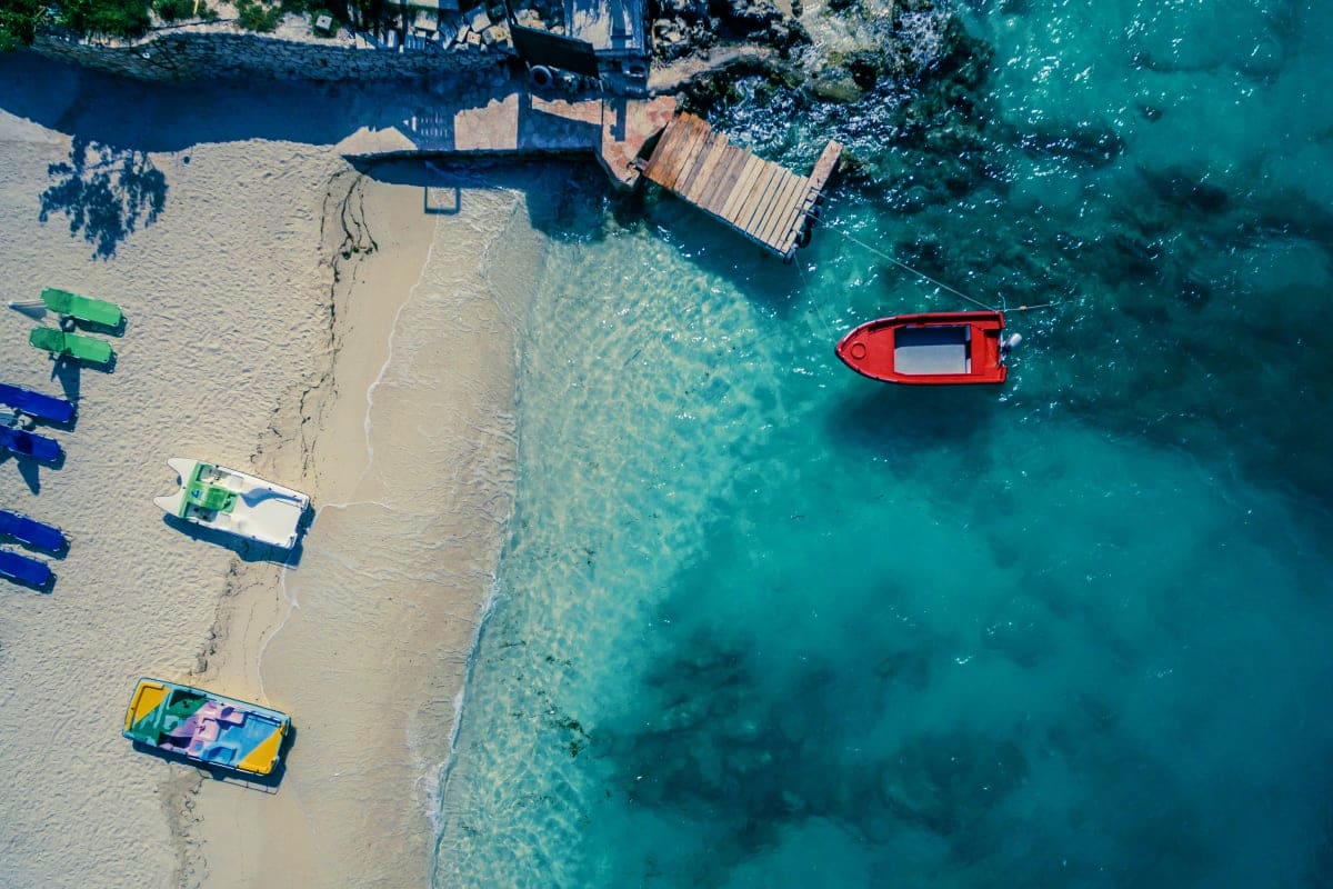 Aerial view of a white sand beach in Albania with colorful fishing boats, wooden pier, and turquoise Mediterranean water