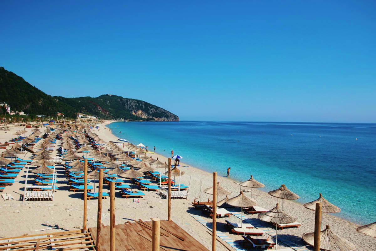 Crowded Mediterranean beach with turquoise water, striped umbrellas, and rocky cliffs in Albania