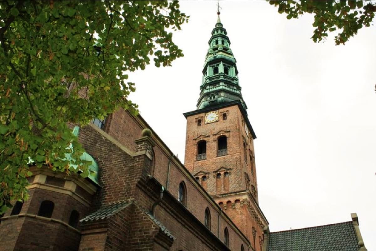 Serbia Luxury Escape Historic brick church tower with green copper spire and clock face, framed by tree foliage in Copenhagen, Denmark