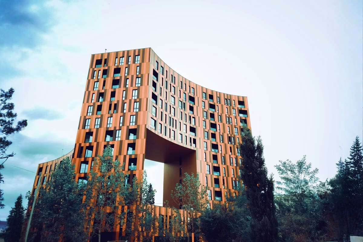 Modern residential building with distinctive curved copper-colored facade and large central archway, surrounded by evergreen 