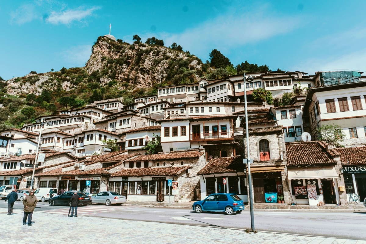 Historic hillside town with white stone buildings cascading up a cliff face, with a blue car and pedestrians in the main squa