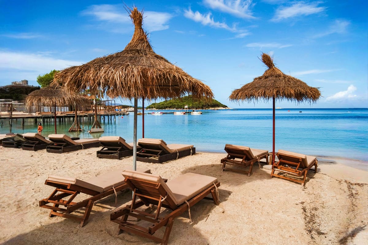 Tropical beach resort with thatched umbrellas, wooden lounge chairs, and turquoise water under clear blue sky