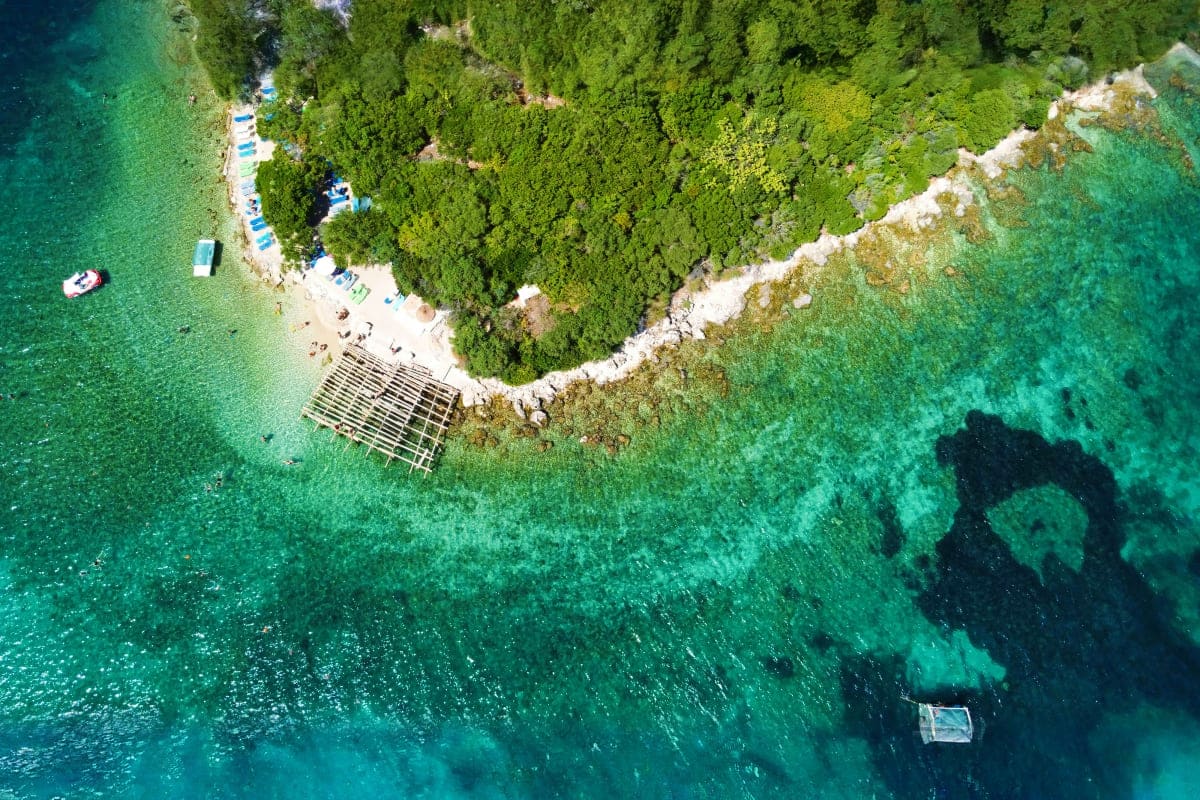 Aerial view of a rocky Adriatic coastline with turquoise water, dense green forest, sandy beach with umbrellas and sunbeds, a