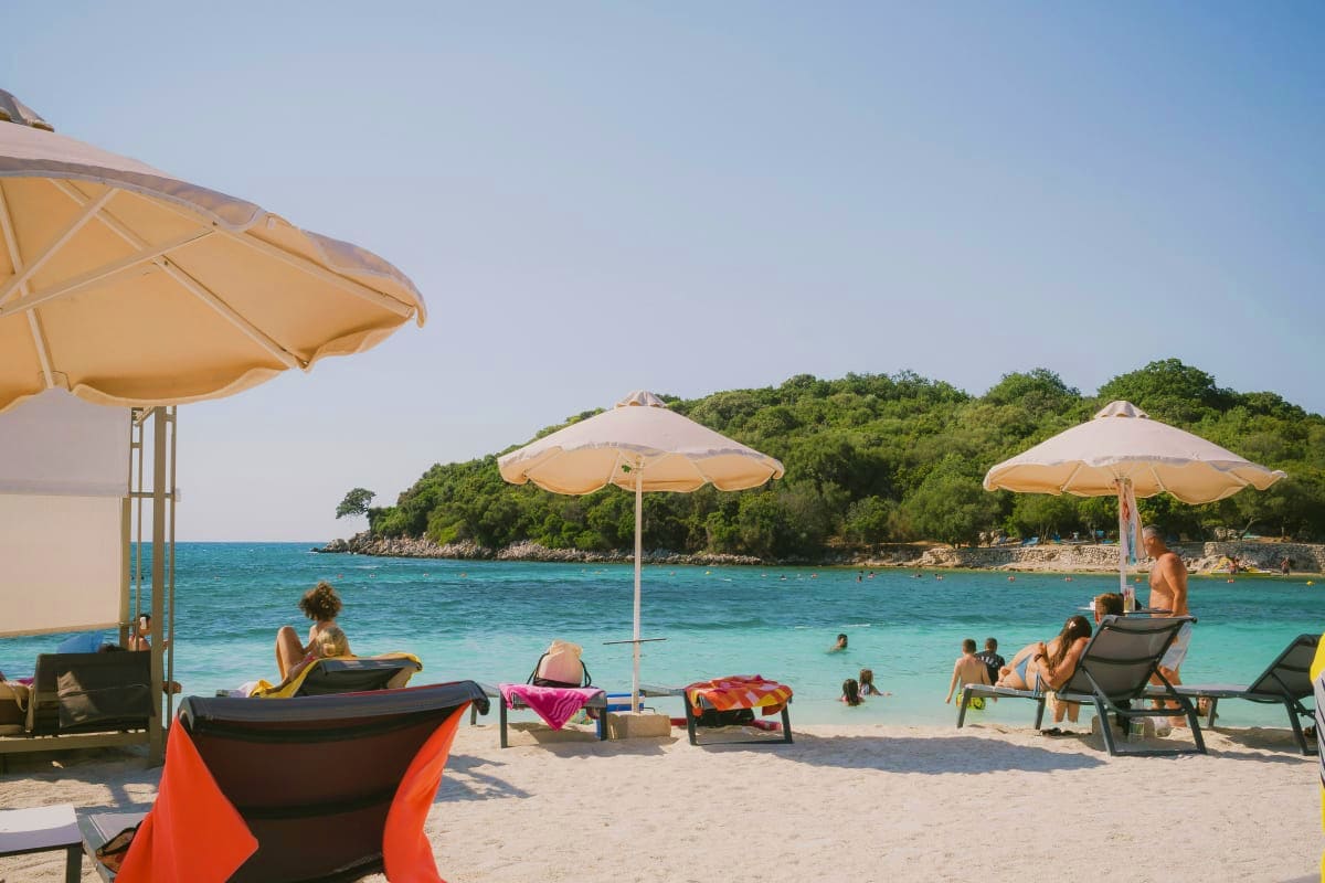 Beachgoers relaxing under cream umbrellas on sandy shore with turquoise Adriatic Sea and forested coastline in background
