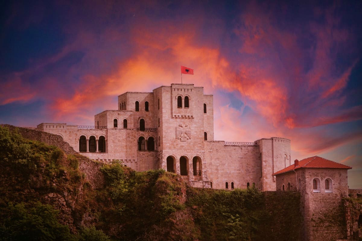 Medieval stone fortress on hillside with Albanian flag at sunset, surrounded by ancient walls and vegetation