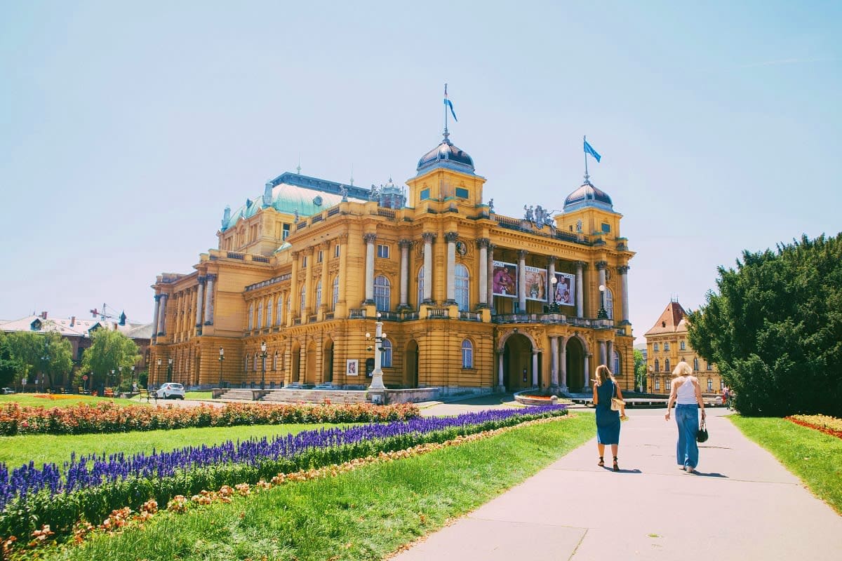 National Theatre building in Zagreb with blue flags, grand yellow neoclassical facade, colorful flower beds, and visitors wal