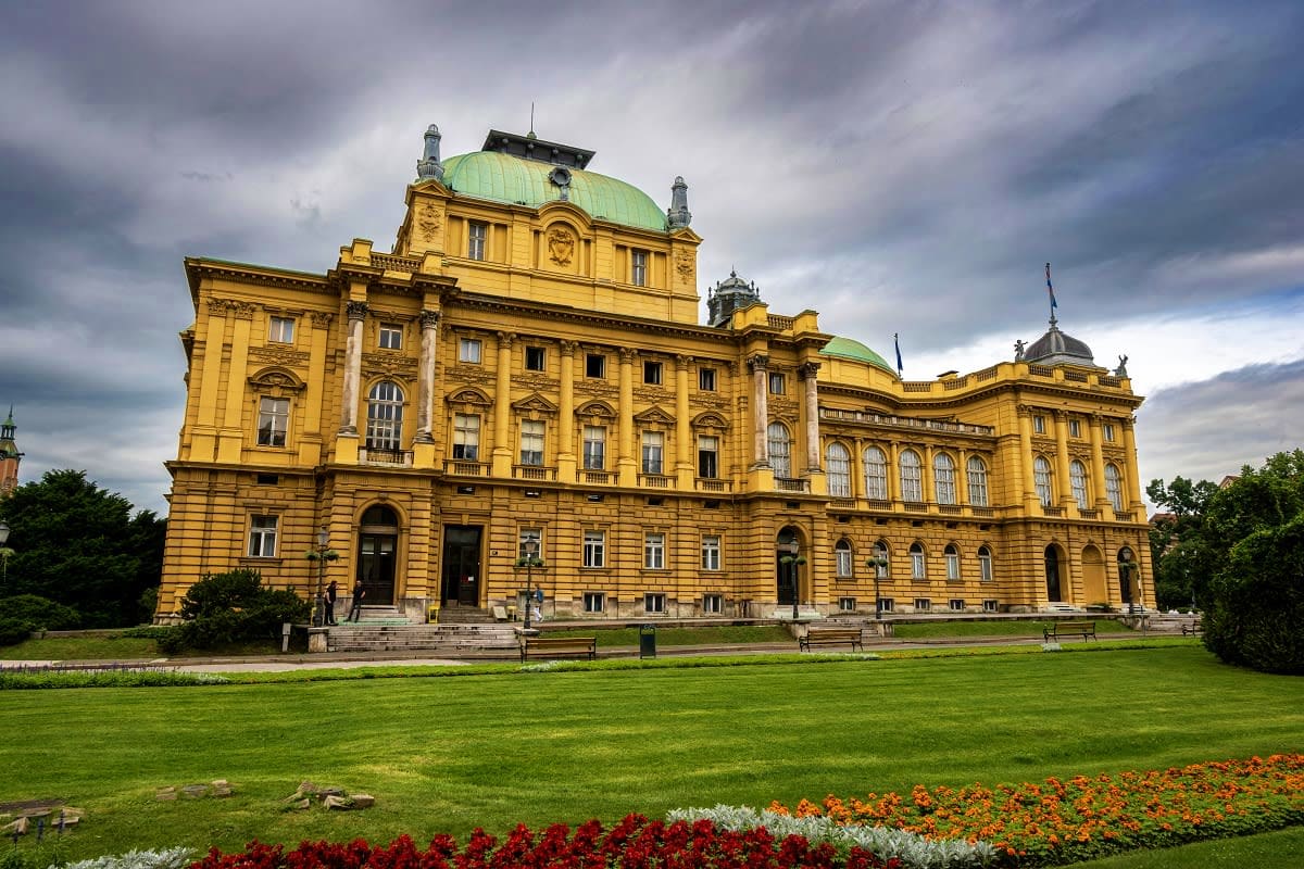 Yellow neoclassical building with green copper domes in Zagreb surrounded by manicured gardens and colorful flower beds